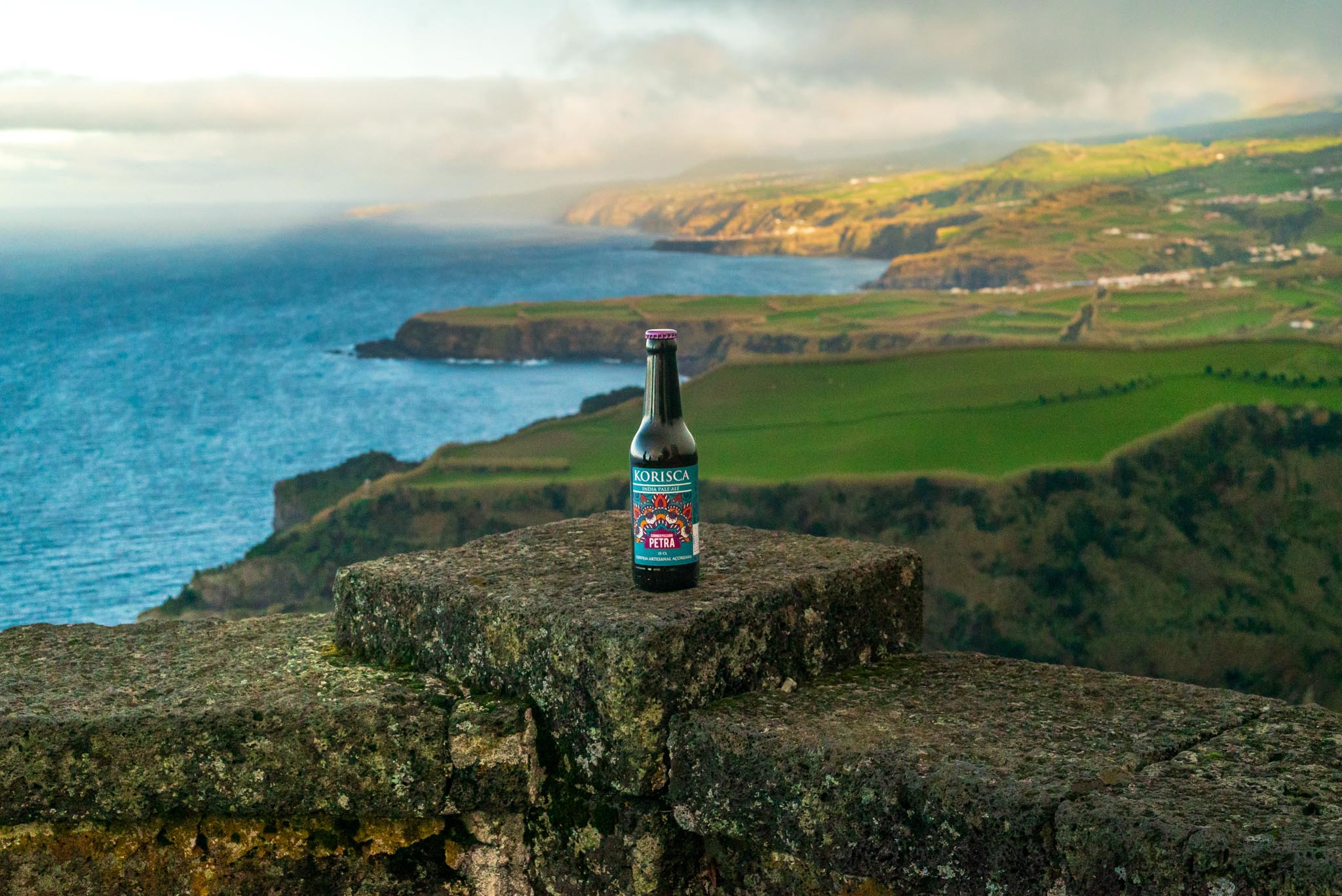 Cerveja artesanal açoriana Korisca Petra (IPA), em cima do muro de pedra com o mar azul, paisagem verde e costa da Ilha de São Miguel de fundo, no Miradouro de Santa Iria, Ribeira Grande, São Miguel, Açores.