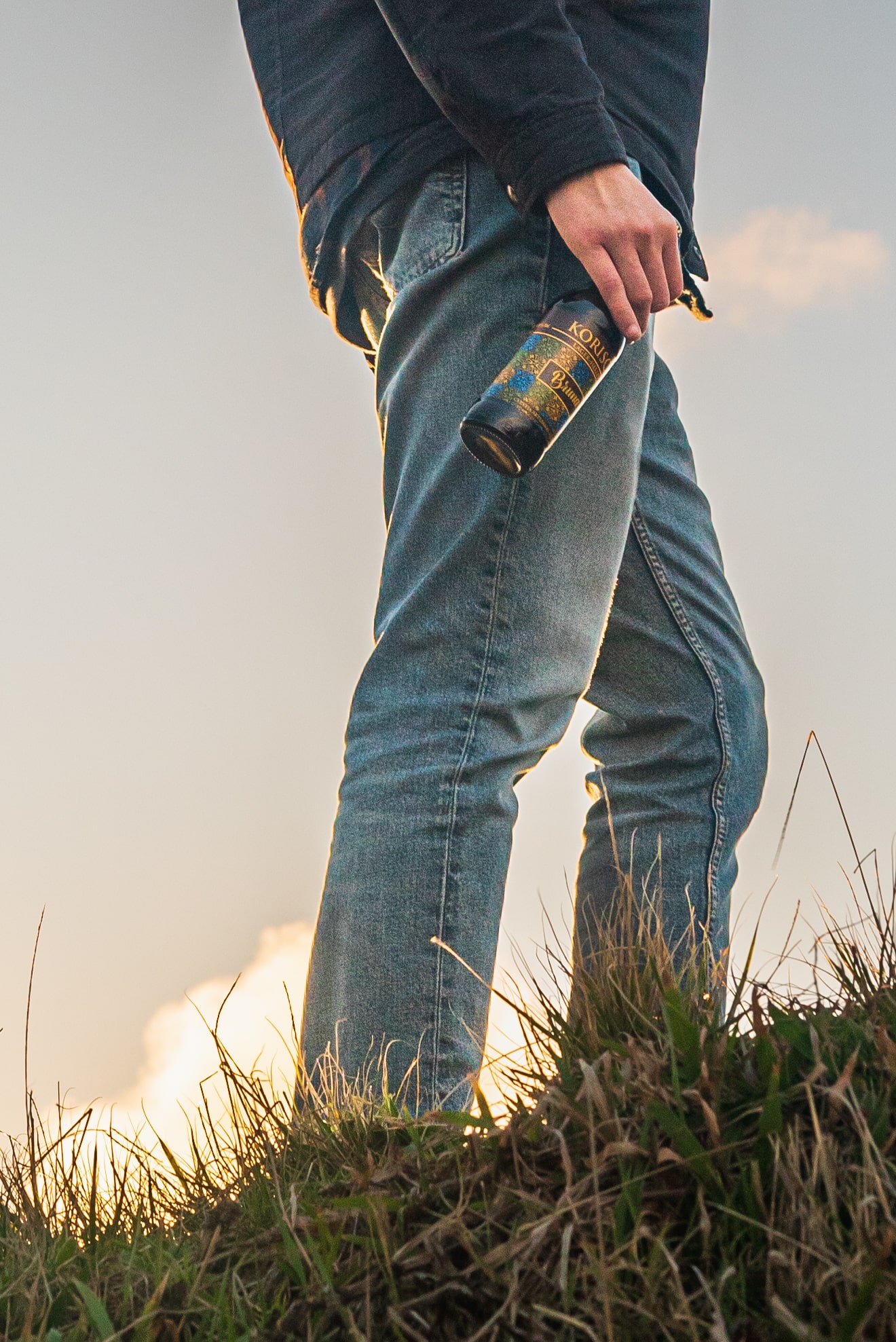 Jovem em cima de vegetação verde, com uma cerveja artesanal açoriana Korisca Bruna (Lager-Helles) na mão e o céu azul com nuvens como fundo, no miradouro da Ponta do Cintrão, na Ribeira Grande, São Miguel, Açores.