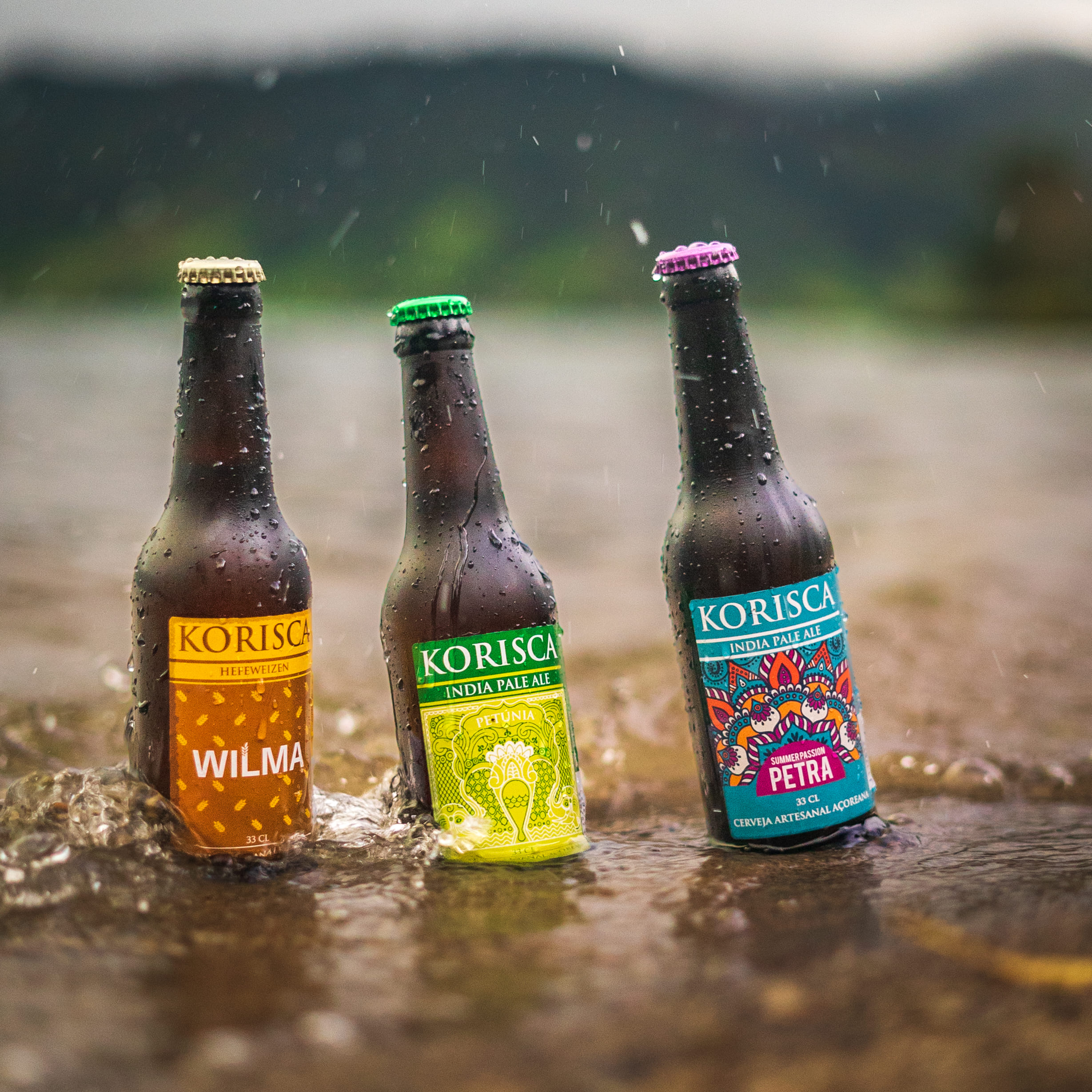 Três cervejas artesanais Korisca, a Wilma (Hefeweizen), a Petúnia (IPA) e a Petra (IPA), na água, com salpicos e ao fundo vale montanhoso, na Lagoa das Sete Cidades, Ponta Delgada, São Miguel, Açores.