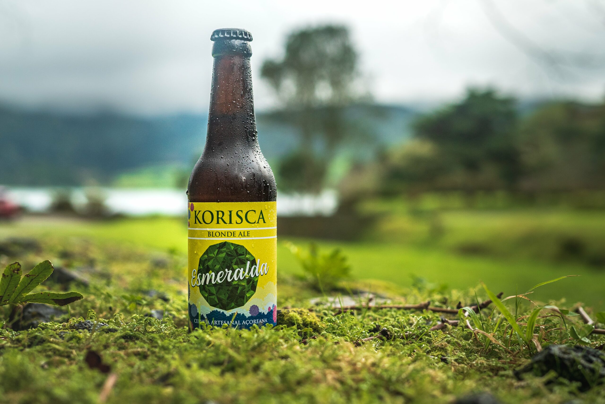 Cerveja artesanal açoriana Korisca Esmeralda (Blonde Ale), em chão verde, com árvores e vegetação verde ao redor, e a Lagoa das Sete Cidades ao fundo, Ponta Delgada, São Miguel, Açores.
