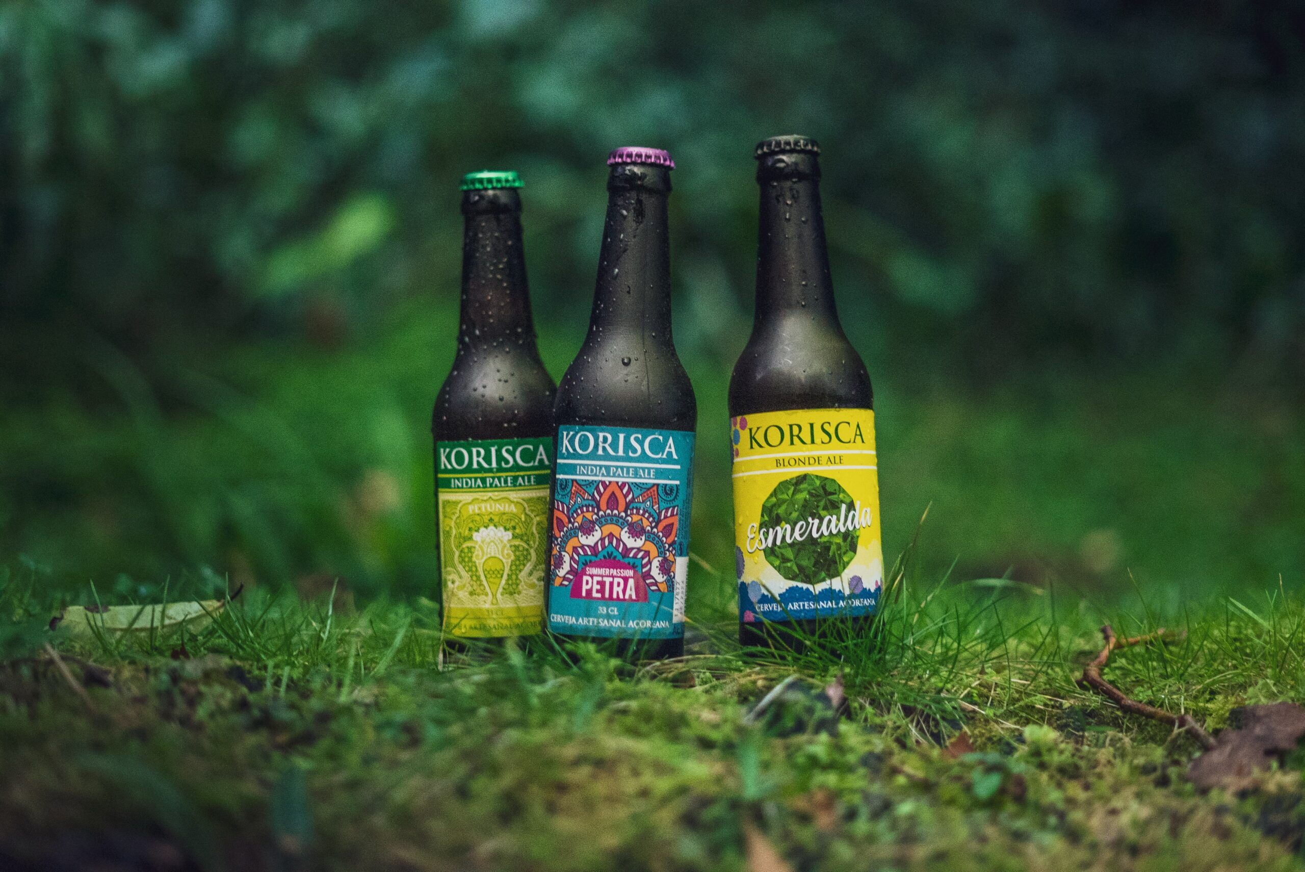 Três cervejas artesanais açorianas Korisca, a Petúnia (IPA), a Petra (IPA) e Esmeralda (Blonde Ale), em chão com erva verde e ao fundo vegetação verde, nas Sete Cidades, Ponta Delgada, São Miguel, Açores.