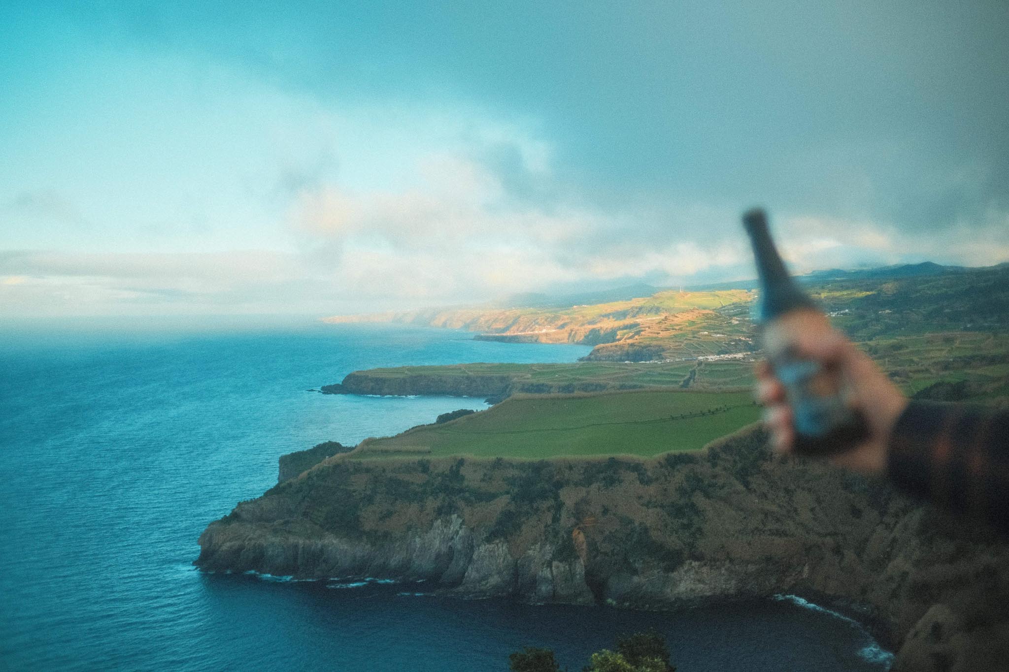 Azorean craft beer Korisca Morgana (Porter) in the hand of a young man, with the blue sea, white clouds and the coast of the island of São Miguel in the background, at the Santa Iria viewpoint, Ribeira Grande, Azores.
