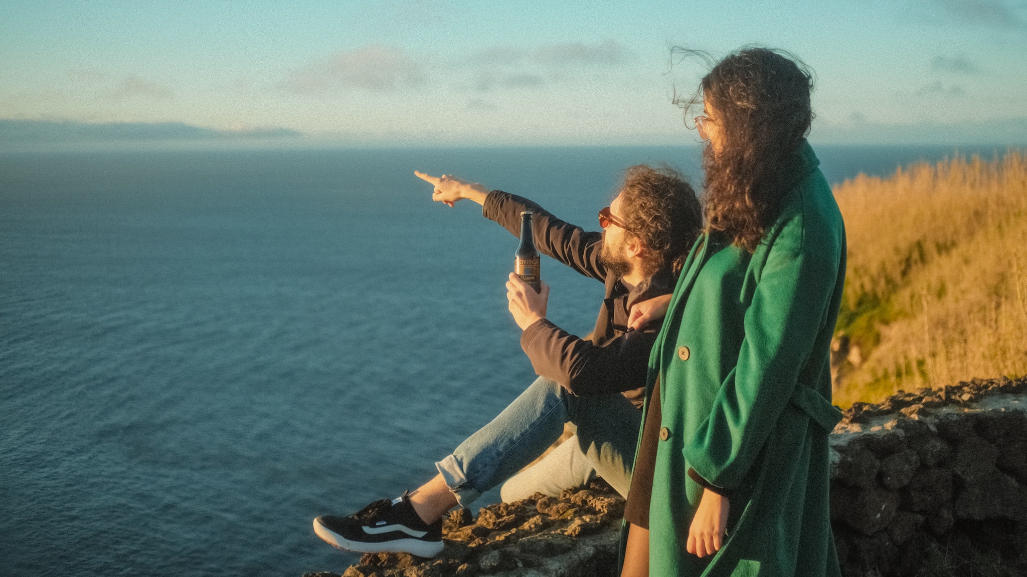 Two young people, on a stone wall, with the blue sea and yellowish vegetation in the background, with the Azorean craft beer Korisca, at the Miradouro da Ponta do Cintrão, in Ribeira Grande, São Miguel, Azores.