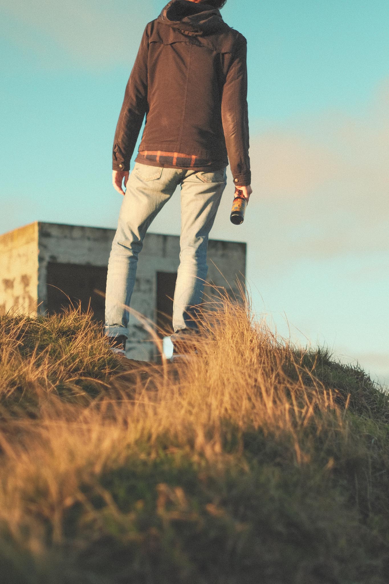 Young man with Korisca Bruna Azorean craft beer (Lager-Helles), on a hill, with a small building (whale watchtower) in the background, and the blue sky with clouds, at the viewpoint of Ponta do Cintrão, in Ribeira Grande, São Miguel, Azores.